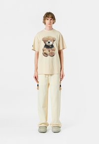 Camiseta beige con un gráfico de osito de peluche, combinada con pantalones crema que tienen borlas. El calzado incluye unos zapatos gris texturizados y sin cordones.