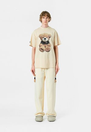 Camiseta beige con un gráfico de osito de peluche, combinada con pantalones crema que tienen borlas. El calzado incluye unos zapatos gris texturizados y sin cordones.