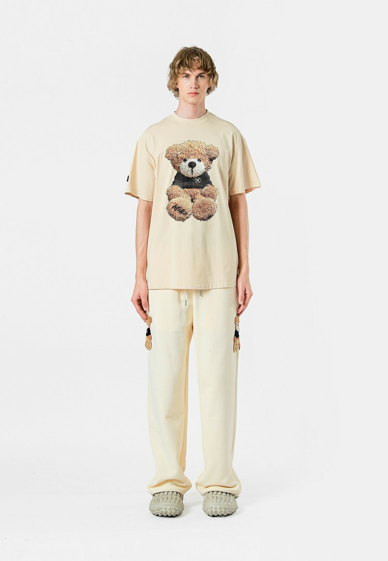 Camiseta beige con un gráfico de osito de peluche, combinada con pantalones crema que tienen borlas. El calzado incluye unos zapatos gris texturizados y sin cordones.