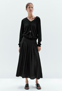 Top nero a maniche lunghe con scollatura a V, abbinato a una gonna nera plissettata lunga fino alla caviglia. Insieme, ci sono semplici sandali neri slip-on e una lunga collana con pendente.