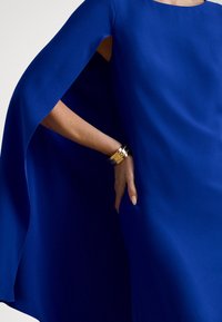 Kobaltkék ruha folyó cape ujjú kialakítással, sima anyagból készült. Arany mandzsettakarkötő látható a csuklón, amely fokozza a megjelenést.