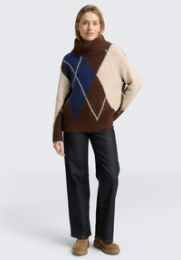 Vrouw staat in een bruine, blauwe en beige argyle coltrui, donkere jeans en tan suède laarzen tegen een effen lichte achtergrond.