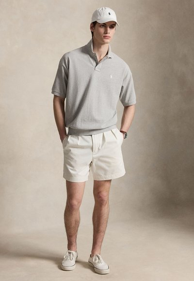 Νέος άνδρας που φοράει ανοιχτό γκρι κοντομάνικο πόλο, λευκά σορτς, λευκό καπέλο και λευκά αθλητικά παπούτσια, στέκεται με τα χέρια στις τσέπες σε ουδέτερο φόντο.
