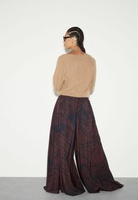 Hnědý pletený svetr s volným střihem, kombinovaný s volnými tmavě hnědými kalhotami s jemným abstraktním vzorem. Model zobrazen zezadu.