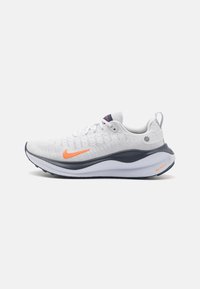 Białe buty sportowe z teksturowaną cholewką, pomarańczowym logo Nike i granatowymi akcentami. Posiadają amortyzowaną podeszwę oraz tylną pętelkę ułatwiającą zakładanie.