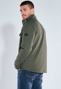 Olivgrüne Jacke mit hohem Kragen, Reißverschluss und vorderer Taschenapplikation. Kombiniert mit blauen Jeans, die eine glatte Textur und eine taillierte Passform aufweisen.