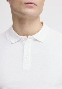 Hvit polo-skjorte med teksturert stoff, klassisk krage, tre-knaps placket og subtil logo på venstre side.