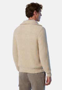 Beige gestrickter Pullover mit weitem Kragen und gerippter Textur, ausgestattet mit einem taillierten Saum. Getragen mit hellen Hosen.