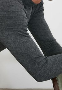 Dunkelgrauer, langärmeliger Strickpullover mit glatter Textur, taillierter Passform und gerippten Bündchen, getragen beim Anlehnen an einen Stuhl.