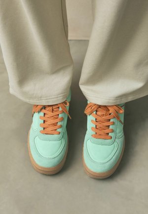 Sneaker in suede verde menta con lacci arancioni e suola in gomma, parzialmente nascosti da pantaloni beige in un contesto neutro.