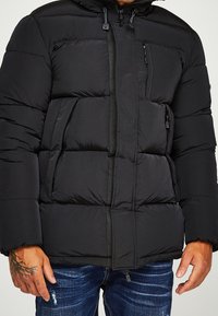 Svart pufferjacka med dragkedja, flera fickor och huva. Tyget ser slätt ut med en quiltad textur.