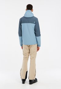 Giacca impermeabile bicolore con cappuccio; corpo azzurro chiaro, parte superiore blu scuro; polsini elasticizzati. Pantaloni beige con aperture con cerniera e accenti neri.