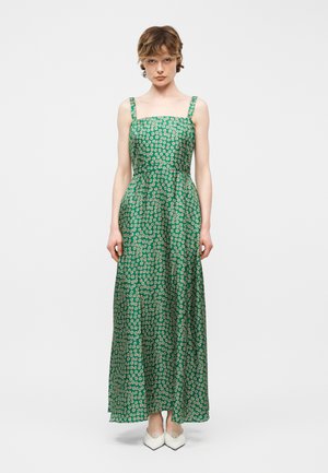 Frau trägt ein grünes ärmelloses Maxikleid mit weißem Blumenmuster und weiße spitze Absatzschuhe, steht vor einem einfarbigen Hintergrund.