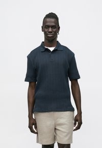 Mørk marineblå strikket poloshirt med korte ermer og krage, med ribbet tekstur og rett kant, kombinert med lyse beige shorts.