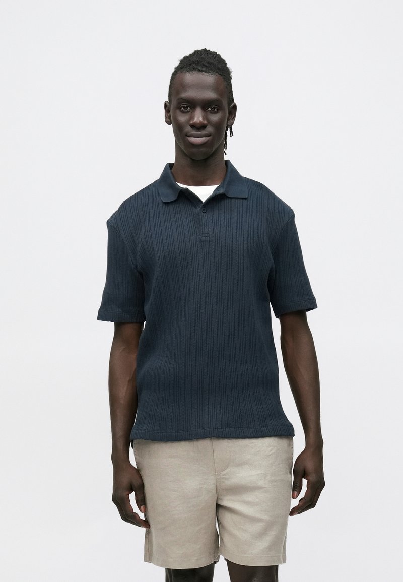 Mørk marineblå strikket poloshirt med korte ermer og krage, med ribbet tekstur og rett kant, kombinert med lyse beige shorts.