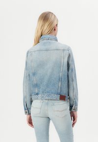 Light blue denim jacket with a collar, front pockets, and a leather patch at the back hem. Styled with matching light blue jeans.