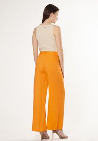 Beige, mouwloos topje met een gestructureerde afwerking, gecombineerd met feloranje, wijde broek met achterzakken, op een model van achteren.