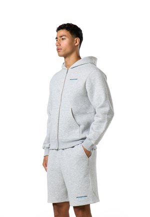 Tânăr purtând hanorac gri deschis cu fermoar și pantaloni scurți asortați, stând cu mâinile în buzunare, uitându-se în lateral.