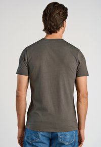 Hombre con pelo corto castaño que lleva una camiseta de manga corta gris oscuro y jeans azules, de pie de espaldas contra un fondo sencillo.