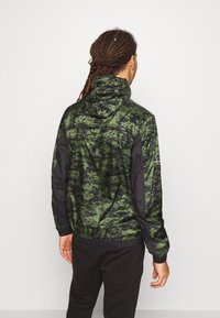 Hættejakke med et grønt og sort digitalt camouflagemønster, med elastiske manchetter, en snoretræk i kanten, og sorte nylonærmer.