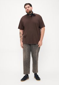 Tricou din bumbac maro cu un croi lejer, asortat cu pantaloni din denim gri decolorat. Adidași negri cu talpă din cauciuc și o bandană neagră la gât.