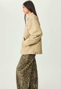 Kvinde med langt mørkt hår iført beige jakke og bredbenede leopardmønstrede bukser, stående i profil mod en ensfarvet baggrund.