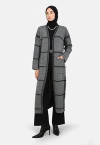 Cardigan lungo grigio con un motivo a quadri, dotato di colletto scialle, due tasche frontali e design aperto sul fronte, abbinato a un abbigliamento nero.