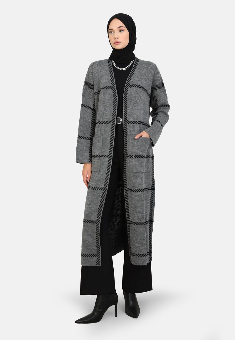Grijze lange cardigan met een geruit patroon, voorzien van een sjaalkraag, twee voorzakken en een open ontwerp, gecombineerd met zwarte kleding.