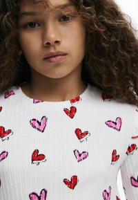 Biely rebrovaný top s motívom srdiečok v červenej a ružovej kontúre. Materiál má hladkú textúru, okrúhly výstrih a žiadne viditeľné ozdoby.