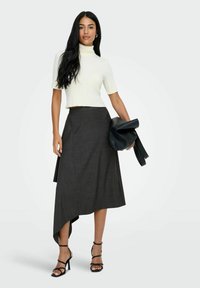 Vrouw in een witte korte mouw turtleneck, asymmetrische donkergrijze rok, zwarte hakken met bandjes, die een zwarte clutch vasthoudt.