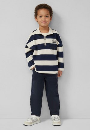 Kind steht, trägt einen marineblau-weiß gestreiften Langarm-Pullover mit Reißverschluss, dunkle Hose und weiße Turnschuhe vor einem schlichten hellen Hintergrund.