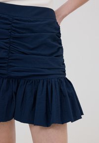 Mörkblå mini-kjol med rynkade detaljer och en utsvängd volangkant. Gjord av lätt tyg och har en texturerad yta.