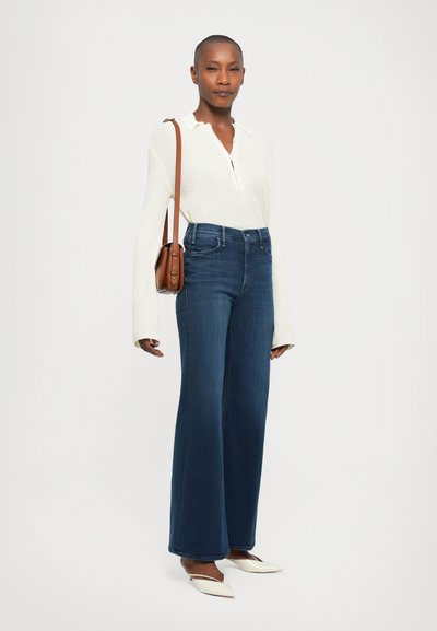 Donna che indossa un top bianco a maglia con maniche lunghe, jeans blu a vita alta e gamba larga, scarpe bianche a punta e una borsa a spalla marrone.