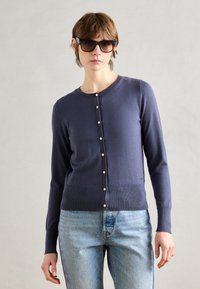 Laivastonsininen neulecardigan, jossa on pyöreä pääntie, ribbireuna ja helmipainikkeet edessä. Yhdistetty vaaleansiniset farkut.