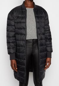 Abrigo acolchado negro con un collar estilo bomber y puños acanalados. Presenta patrones de acolchado horizontal y un cierre frontal completo.