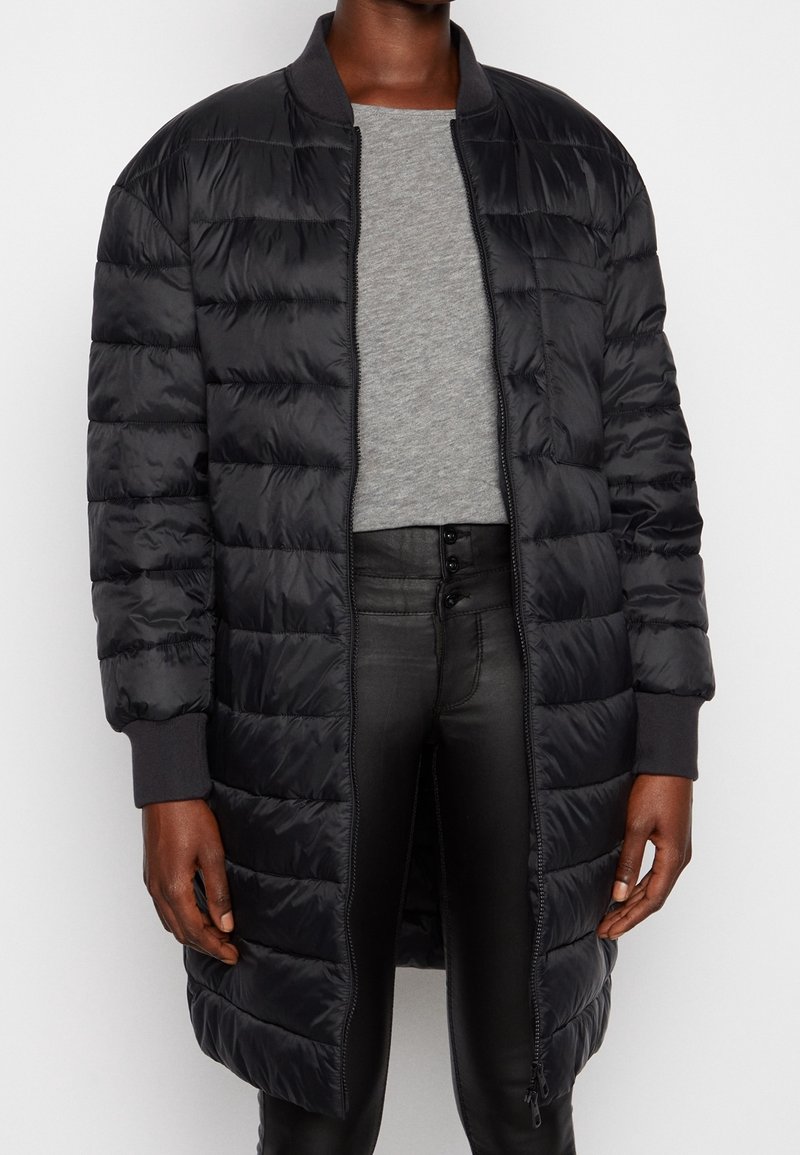 Abrigo acolchado negro con un collar estilo bomber y puños acanalados. Presenta patrones de acolchado horizontal y un cierre frontal completo.