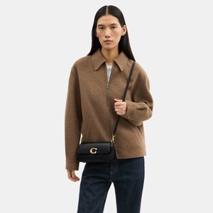 Žena nosiaca hnedú bundu na zips a tmavé džínsy, nesúca čiernu kabelku cez telo so zlatou prackou na bielom pozadí.