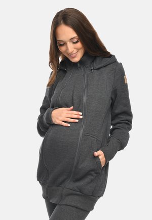 Schwangere Frau in dunkelgrauem Kapuzenpullover mit Reißverschluss, die die Hand auf den Bauch legt, lächelt und nach unten schaut, vor einem einfarbigen Hintergrund.