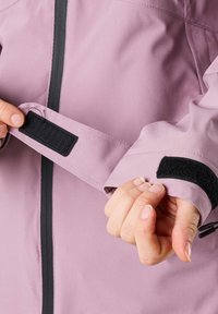 Ljuslila jacka ärmslut med svart kardborreband. Materialet har en slät textur. Dragkedjestängning och justerbara funktioner är synliga.