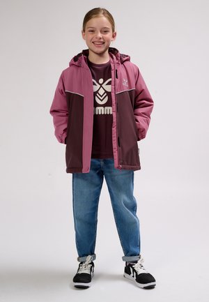 Smilende barn står med hænderne i lommerne iført en lilla tofarvet jakke, en burgunder t-shirt med tryk, blå jeans og sorte-hvide sneakers.