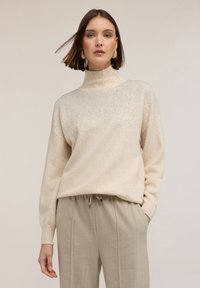 Kobieta ubrana w beżowy sweter z wysokim kołnierzem, zdobiony subtelnym brokatem, oraz luźne beżowe spodnie z trokami na tle neutralnym.