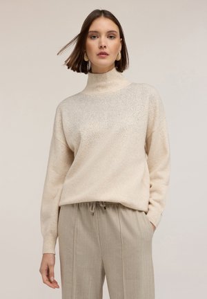 Kobieta ubrana w beżowy sweter z wysokim kołnierzem, zdobiony subtelnym brokatem, oraz luźne beżowe spodnie z trokami na tle neutralnym.