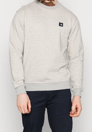 Personne portant un sweat-shirt gris clair à col rond avec un petit patch logo noir sur la poitrine gauche, assorti à un pantalon bleu marine foncé.