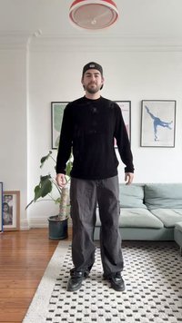 Black long-sleeve shirt and gray cargo pants worn by a person standing on a patterned rug near a plant and a light fixture.