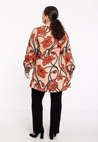Blomster tunika i beige med røde blomster og sorte kantlinjer, forsynet med lange puffærmer og en buet søm, parret med sorte bukser.