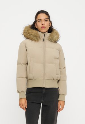 Mujer con chaqueta acolchada beige con capucha con ribete de piel y pantalones negros, de pie frente a un fondo blanco liso.