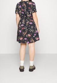 Schwarzes Blumenkleid mit kurzen Ärmeln und tailliertem Schnitt, verziert mit lila und pinken Blumenmustern. Getragen mit weißen Socken und grünen Schuhen.