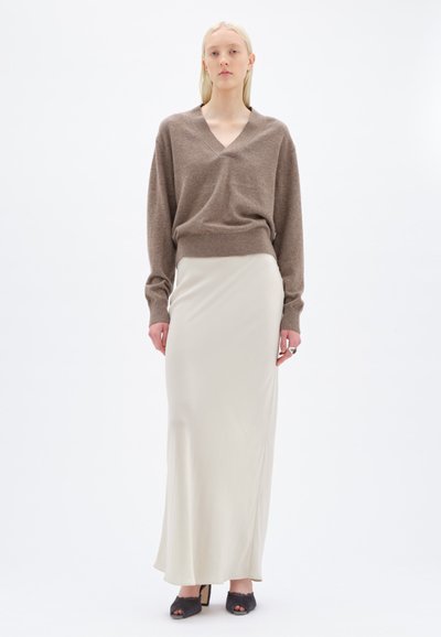 Maglione marrone a collo a V con vestibilità morbida, abbinato a una lunga gonna in satin beige chiaro. La modella indossa tacchi aperti neri e un anello.