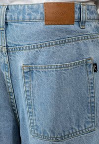 Jeans de mezclilla azul claro con un parche de cuero y dos bolsillos traseros, que presentan costuras en contraste y una ligera textura desteñida.