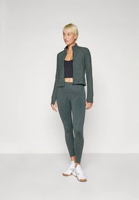 Set format din geacă sport verde închis și colanți, confecționat dintr-un material texturat cu un design ajustat, asortat cu un top scurt negru și adidași albi.